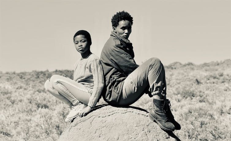 A black and white photo of an African girl and an African woman sitting back to back on a rock, looking at camera.