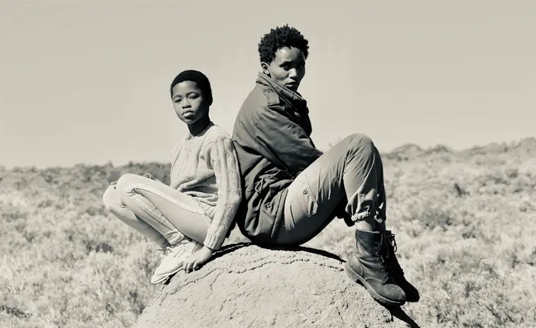 A black and white photo of an African girl and an African woman sitting back to back on a rock, looking at camera.