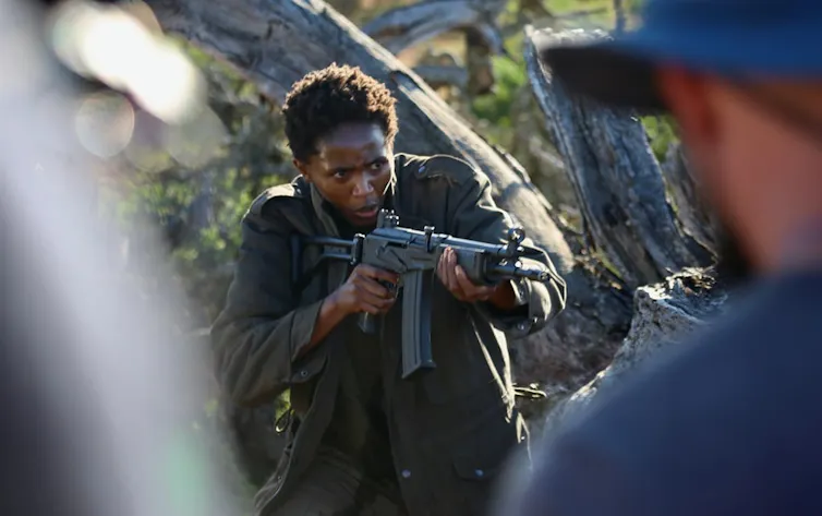 An African woman in military-style khaki clothing holds a rifle, poin it at a man wearing blue.