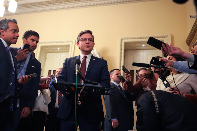 Man in suit and tie speaks at a podium surrounded by the media and more men in suits and ties.