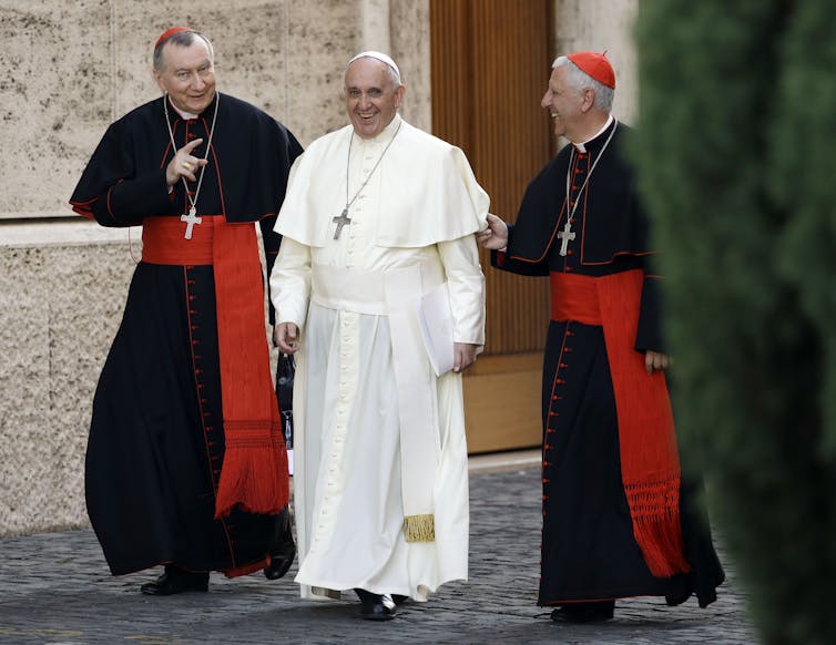 Two men in black robes with red skullcaps and red sashes walk on a paved road, flanking a man in white robes.