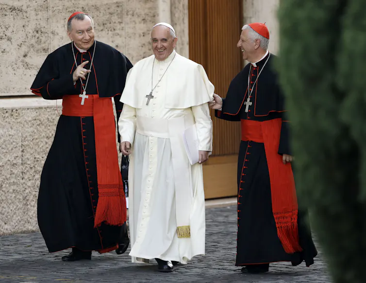 Two men in black robes with red skullcaps and red sashes walk on a paved road, flanking a man in white robes.