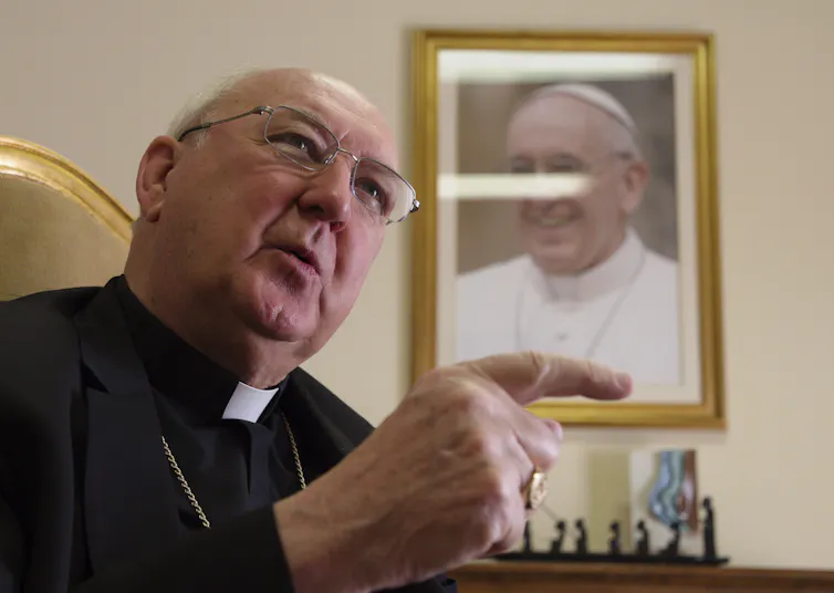 A man wearing a priest's collar gestures as he speaks, sitting in front of a framed portrait of Pope Francis.