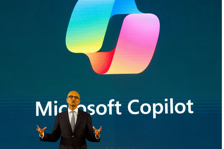 Photo of a man in a suit talking in front of a screen showing the Microsoft Copilot logo.