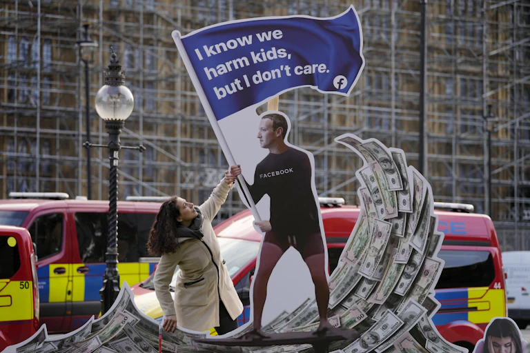 A cardboard cutout of Mark Zuckerberg riding a wave of cash, carrying a blue flag that says: "I know we harm kids, but I don't care."
