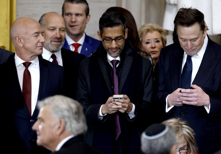 A bald, besuited man looking over at two other besuited men on their phones.