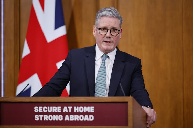 British Prime Minister Keir Starmer wearing a dark suit and grey tie standing at a podium framed by the Union Jack