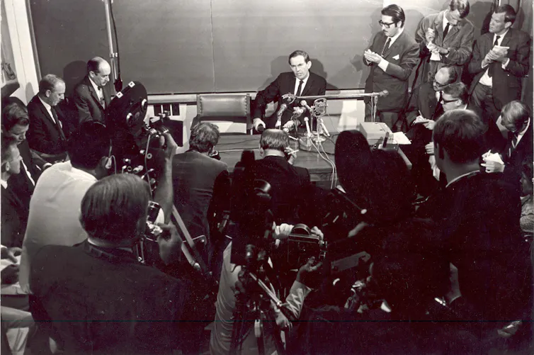 A black and white photo of a man sitting at a table surrounded by other men with cameras and notepads