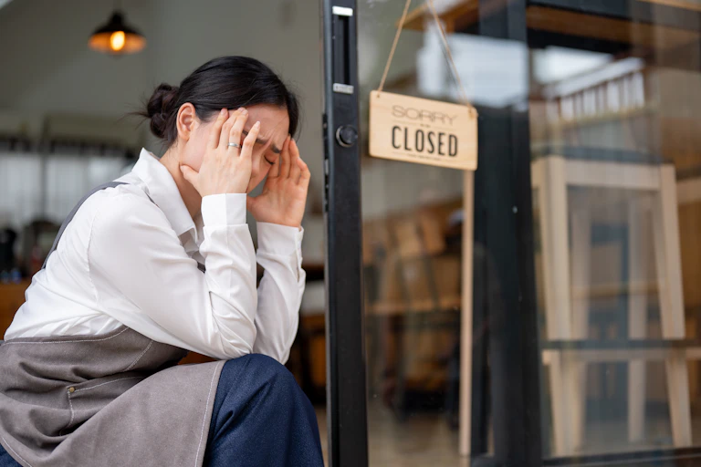 woman with her head in her hands outside her business with a 'closed' sign on the door.