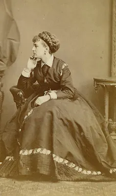 An actor sat in a chair dressed in a woman's long dress with hair tied back.