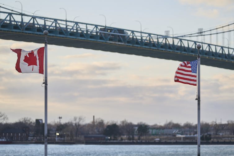 Two flags fly in front of a vehicular bridge.