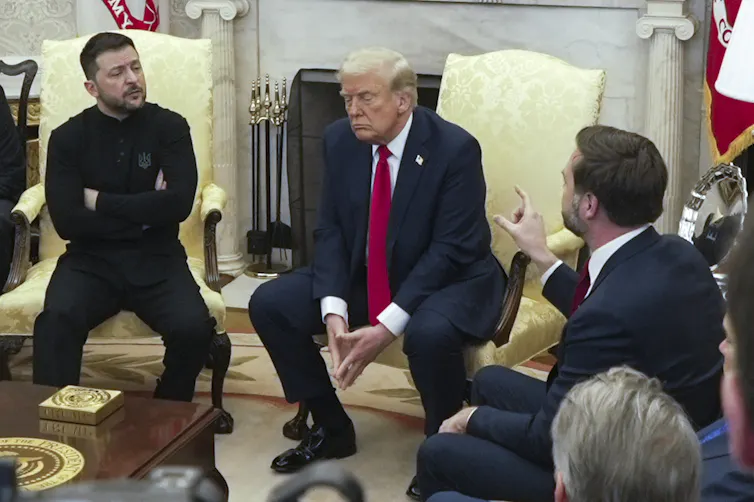 Three men sit in discussion, one gesturing with his hand pointing upwards.