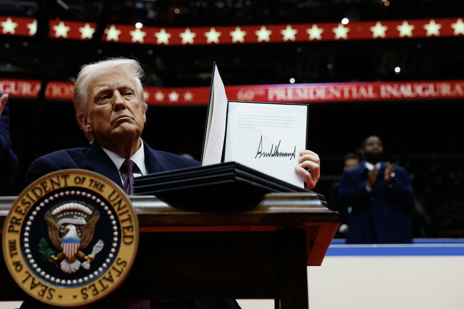 Donald Trump holds a signed executive order behind a table or a lectern bearing the US presidential seal at an indoor event. 