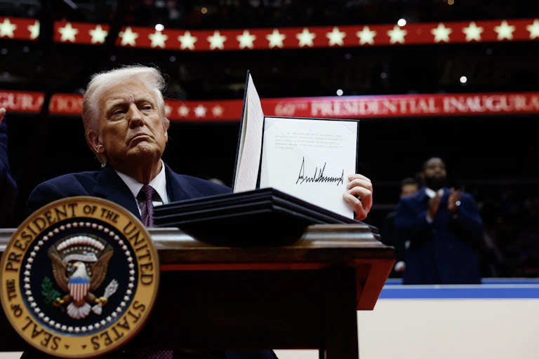 Donald Trump holds a signed executive order behind a table or a lectern bearing the US presidential seal at an indoor event.