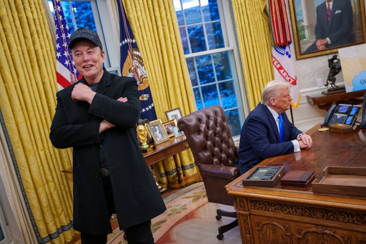 A man stands while behind him a man sits at a desk.