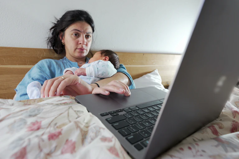 An exhausted woman holding a newborn uses a laptop while sitting up in a bed