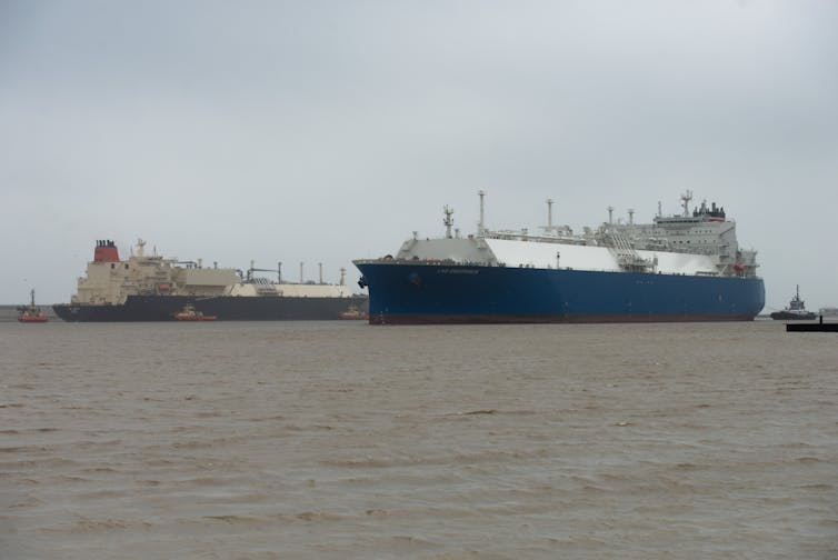 One large ship moves in front of another large ship; both are attended by tugboats.