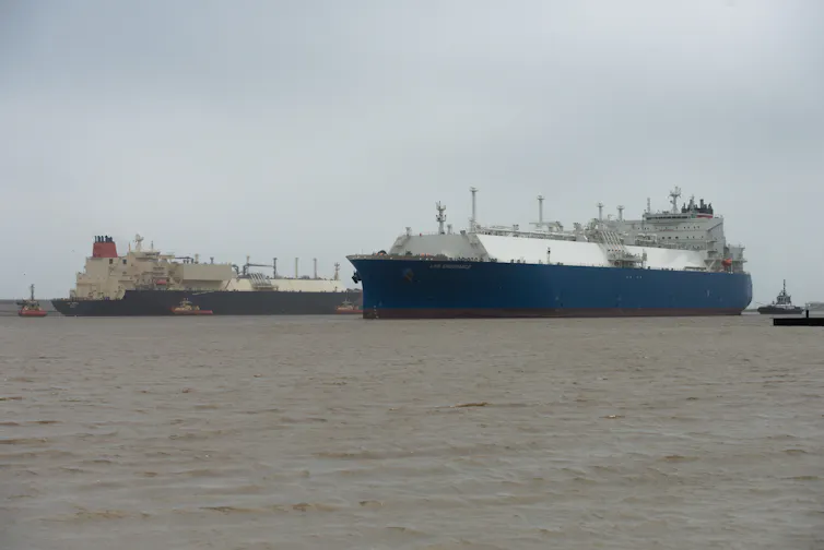One large ship moves in front of another large ship; both are attended by tugboats.