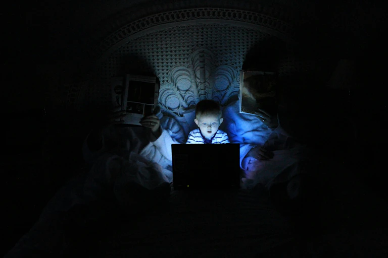 A young boy looking at a computer screen in bed, next to two adults.