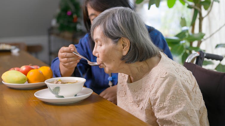 A middle-aged woman feeds soup to an older woman, both are seated at a dining table