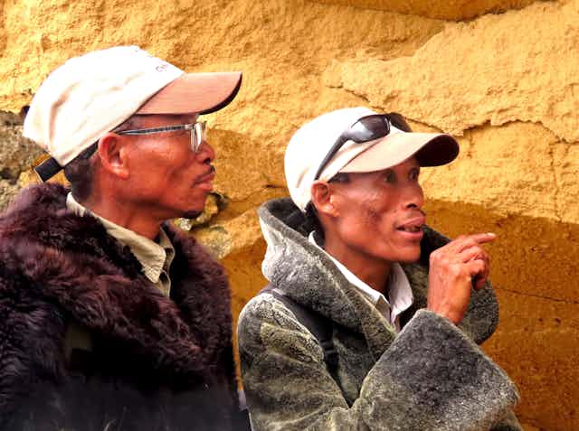 Two men wearing caps and warm jackets stand in front of a rocky wall. One of them is pointing ahead.