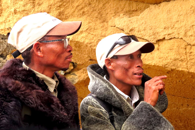 Two men wearing caps and warm jackets stand in front of a rocky wall. One of them is pointing ahead.