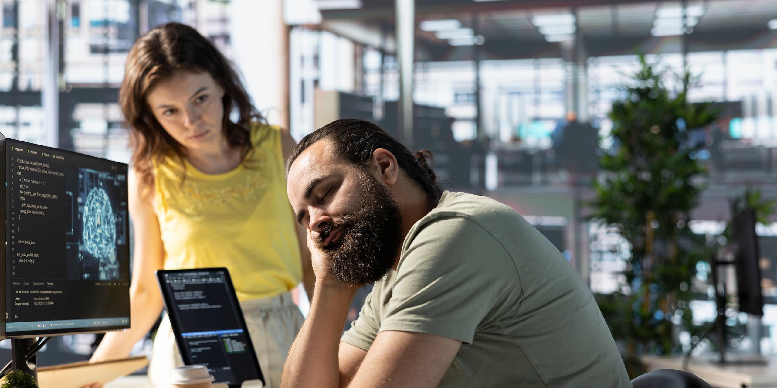 A woman in a yellow top looks on as a man in a light green t-shirt props his head in his hand and has his eyes closed at a desk in front of a computer screen.