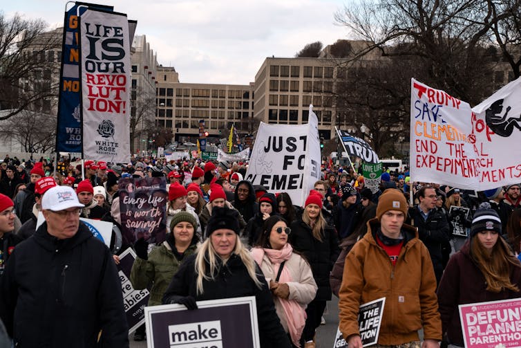A large crowd marches while holding banners with various slogans. One such banner reads, 'Life is our Revolution.'