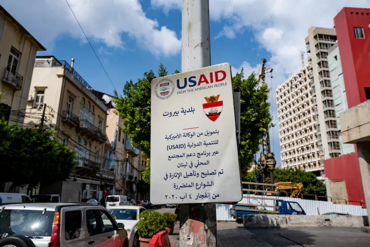 A plaque on a poll says USAID and has writing in Arabic on a busy city street.