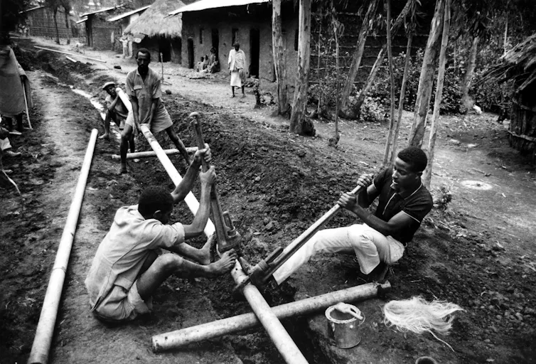 People work on a long-exposed metal pipe in a rural-looking town in a black-and-white photo.