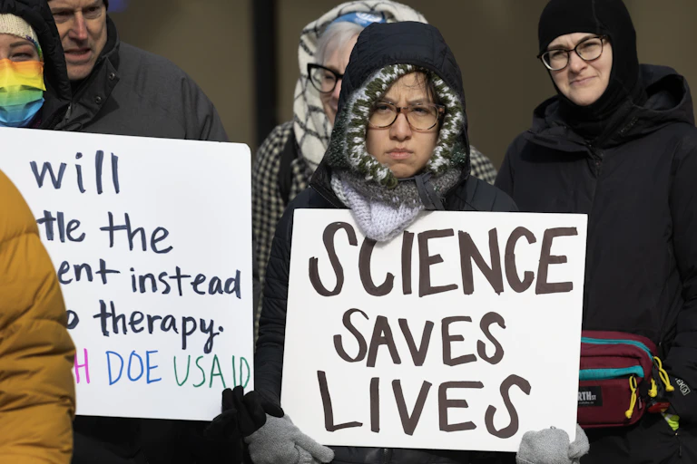 A protestor wearing a heavy jacket holds up a sign that says "Science Saves Lives."