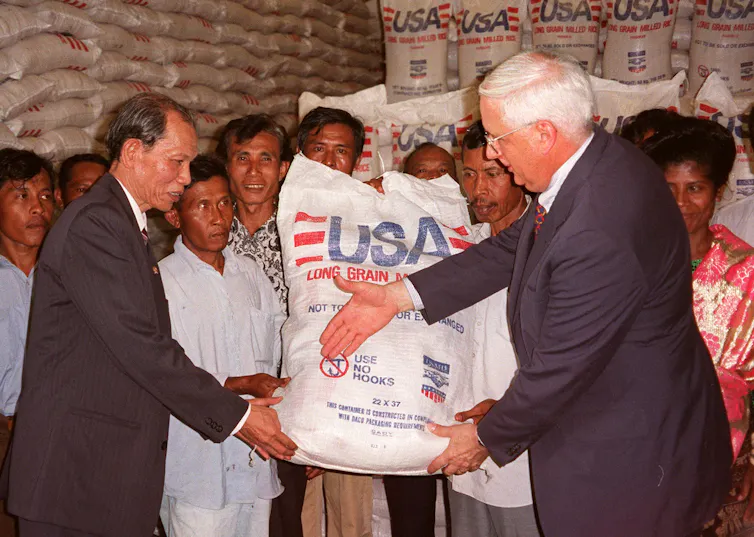 Two men wearing suits appear to move to shake hands, with people standing behind them carrying a large white bag that says USAID.