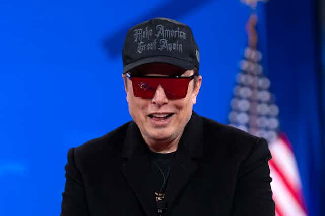 a smiling man wearing a dark jacket over a T-shirt, dark ball cap and sunglasses