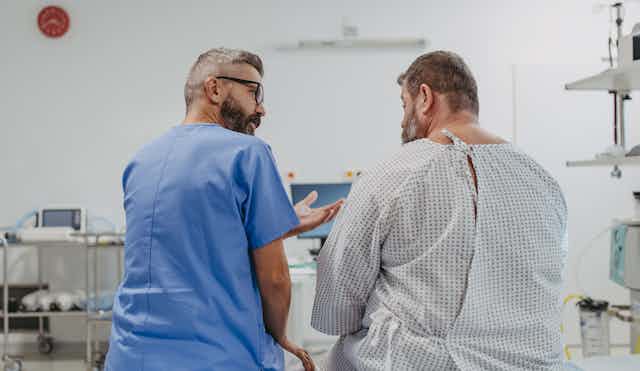 A doctor in blue scrubs sitting with an overweight man in a medical gown, seem from behind