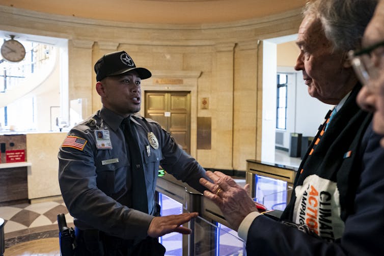 a security guard holds out his arms as he stands in front of an older man in a business suit