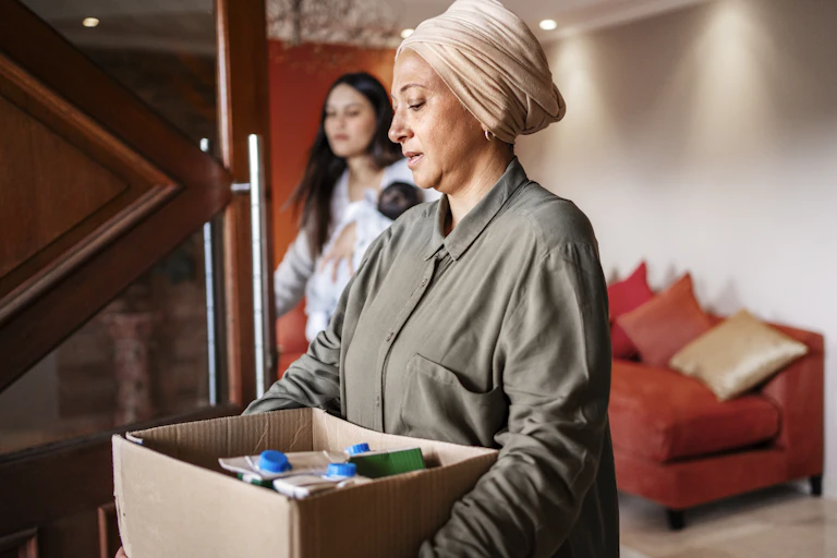 A woman wearing a headdress carries a box containing cartons of food, while a younger woman walks beside her.
