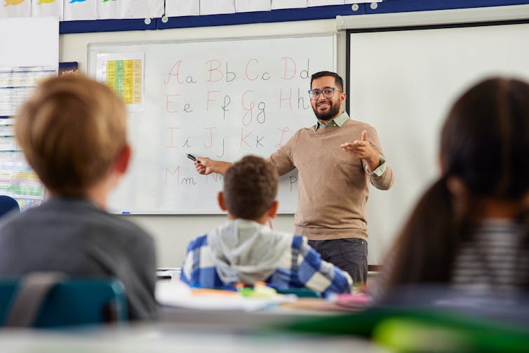 Male teacher writing on a whiteboard in front a group of children.