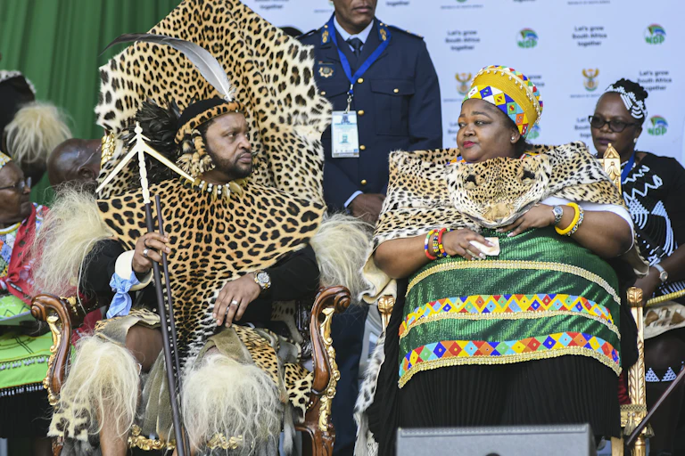 An African man and woman sit looking at one another with frank expressions. In gilded chairs they are dressed in full traditional Zulu finery with leopard skins and beaded fabrics.