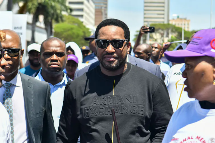 A bearded African man in shades and a black Armani sweatshirt walks with a group of men down a street.