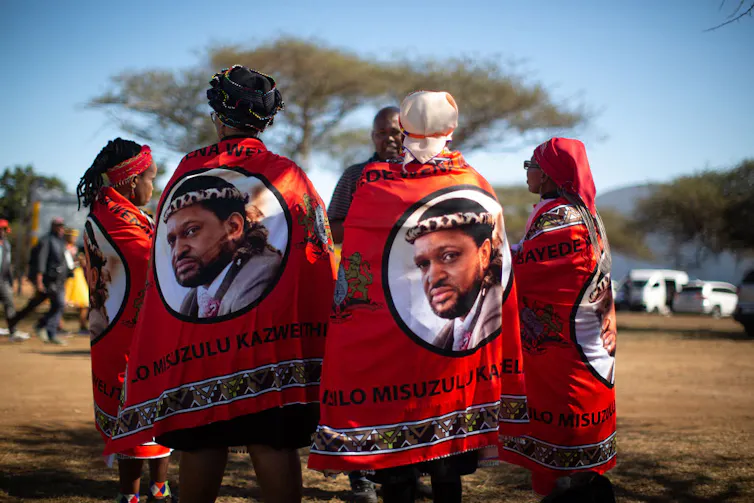 Four African women stand, their backs to camera in a rural setting. They are draped in bright red cloths that bear the image of a bearded African man.