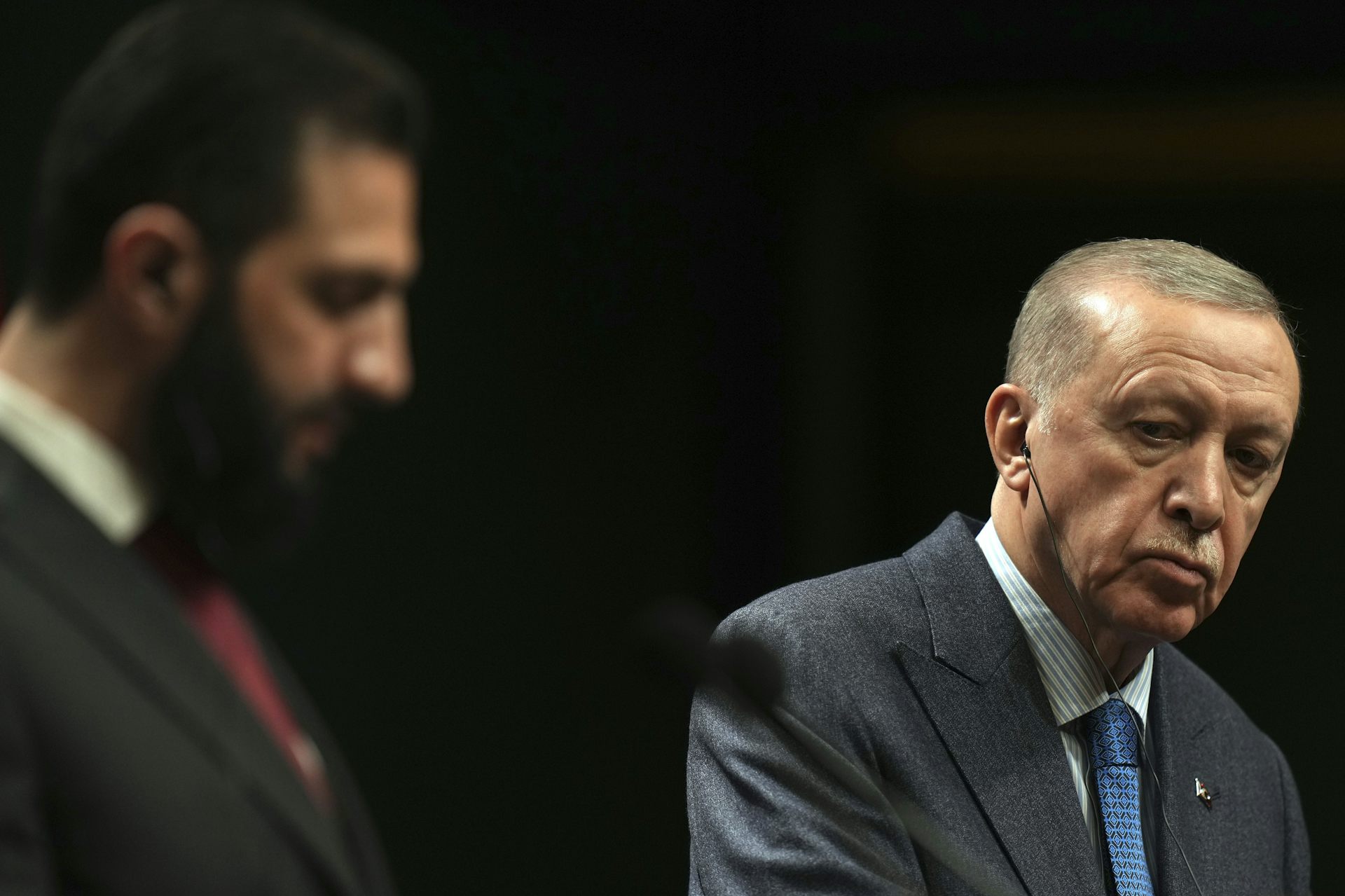 A man with thin grey hair listens while a bearded man, out of focus, speaks.