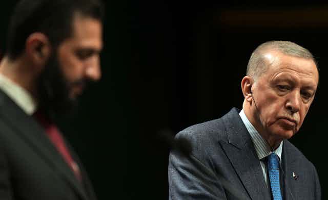 A man with thin grey hair listens while a bearded man, out of focus, speaks.