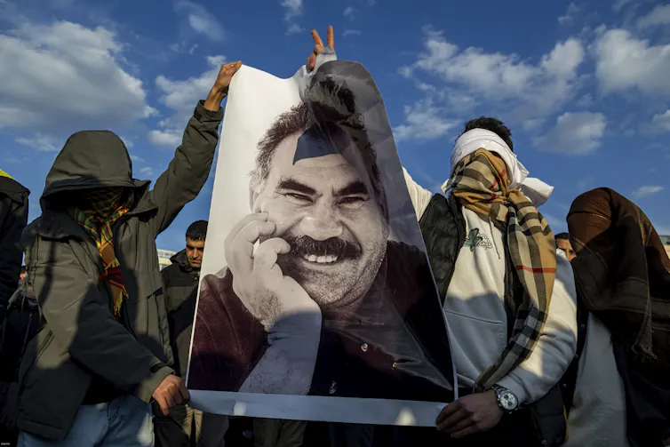 Young people with with scarves over their faces hold up a poster of a smiling man with a moustache.