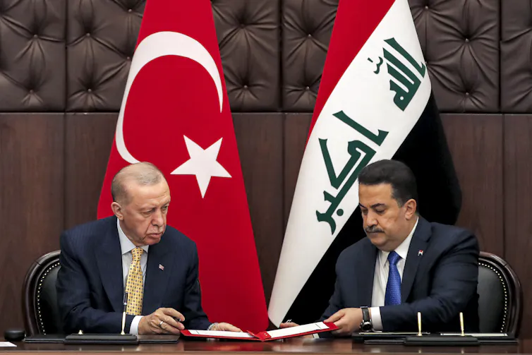 Two men, one with thinning grey hair and a younger man with black hair and a moustache, sit at a table and look at documents.