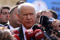 An older bald man surrounded by microphones.
