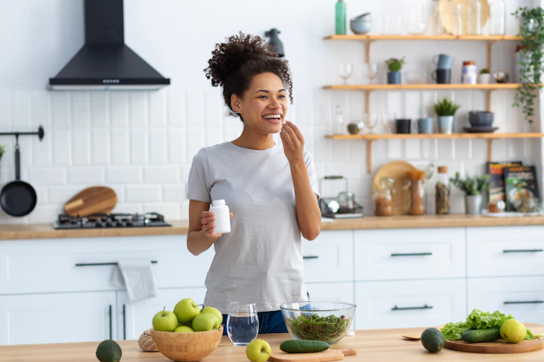Woman standing in kitchen taking a health supplement, green fruit and vegetables are on the kitchen counter in front of her