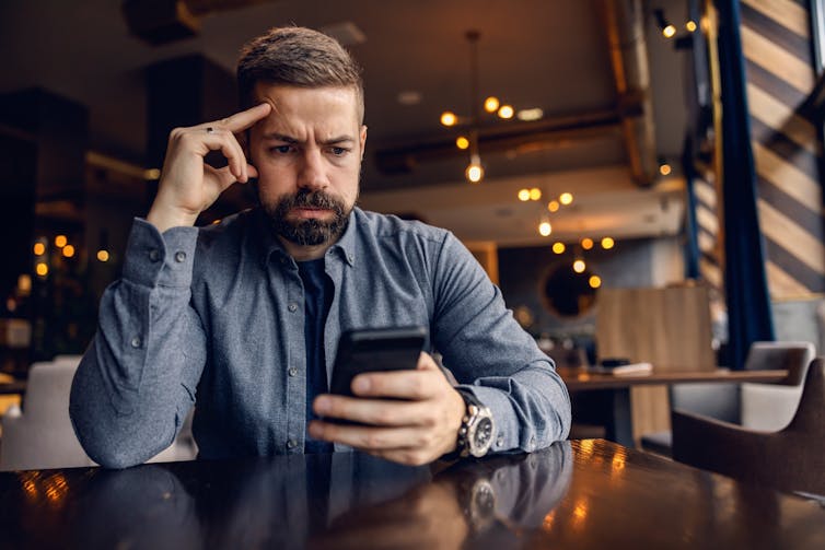 A man sitting in a cafe using a phone with a worried expression on his face