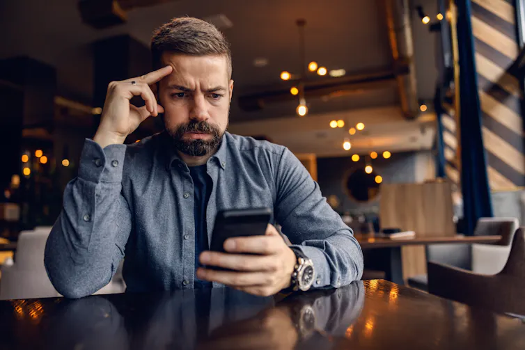 A man sitting in a cafe using a phone with a worried expression on his face