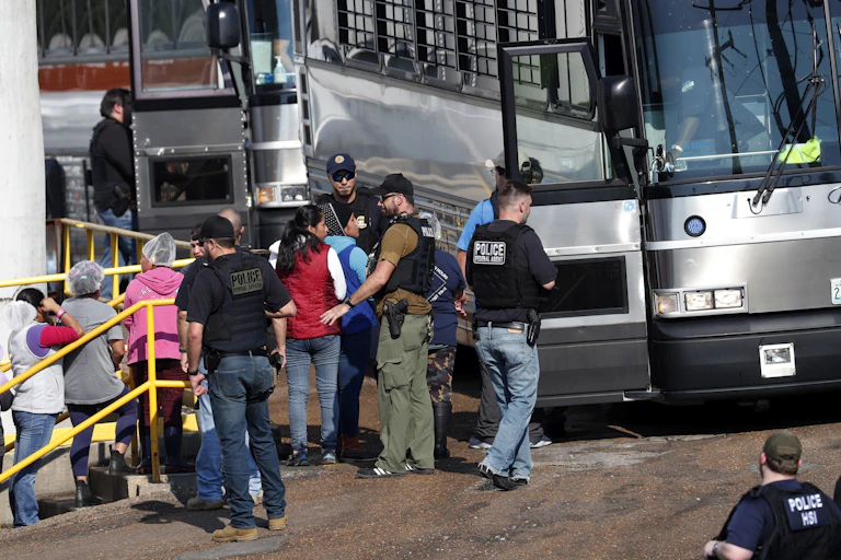 Men with guns make women, some in hairnets, line up and board a bus with bars on its windows.