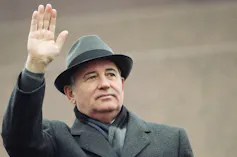 A man in a fedora and overcoat waves.